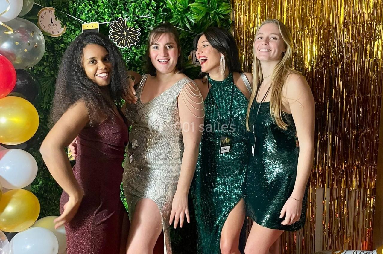 Quatre femmes en robes de soirée posent souriantes devant une décoration festive avec ballons et rideau doré.