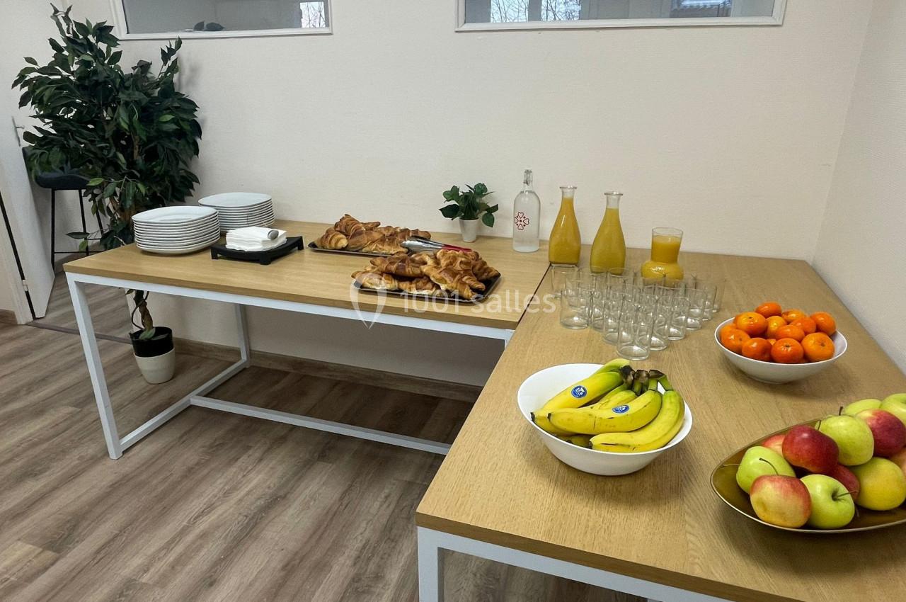 Buffet avec fruits, viennoiseries, jus d'orange et vaisselle disposés sur deux tables en bois dans une pièce lumineuse.