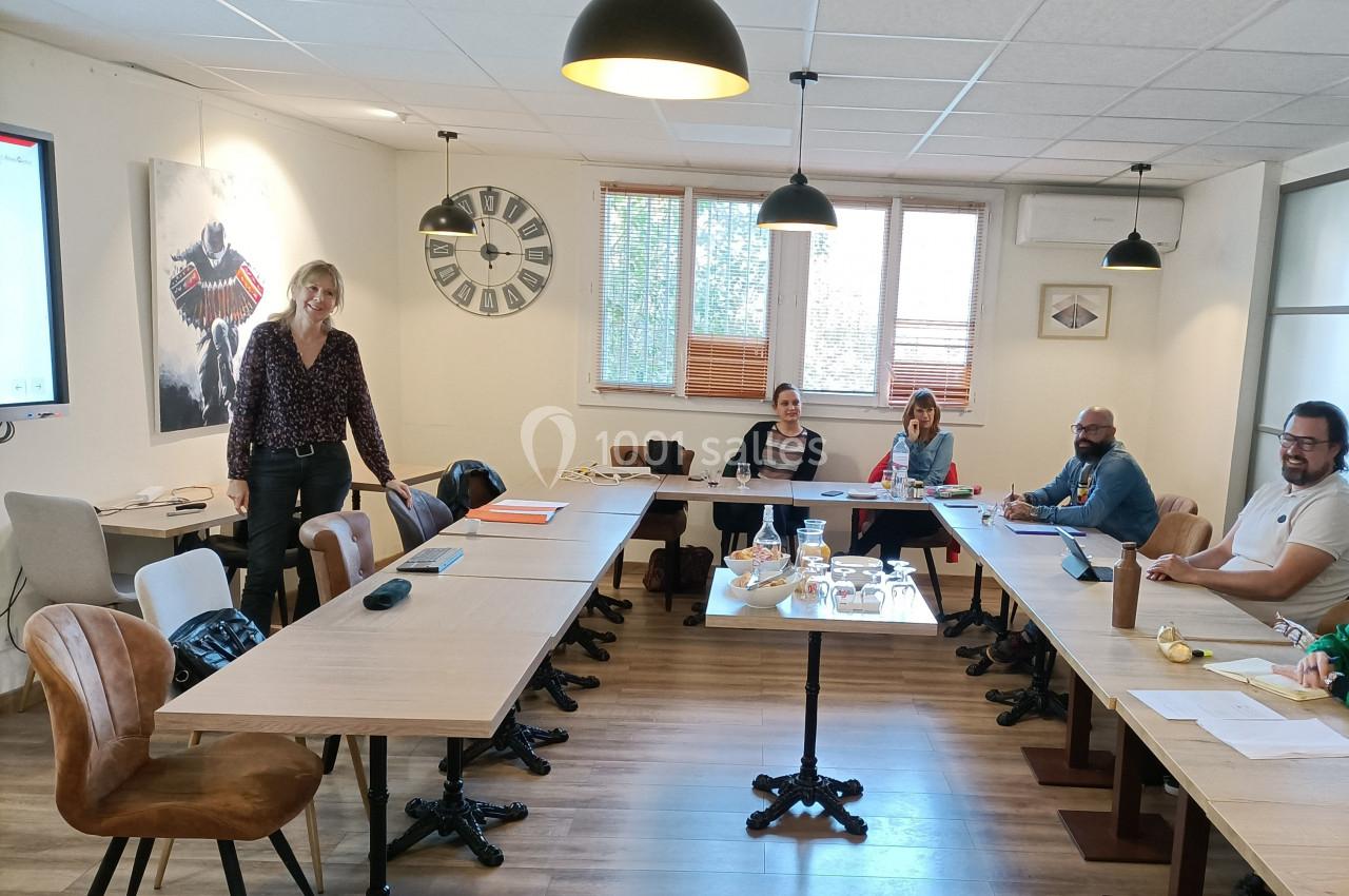 Une salle de réunion avec des personnes assises autour de tables en bois, une femme debout près d'un écran.