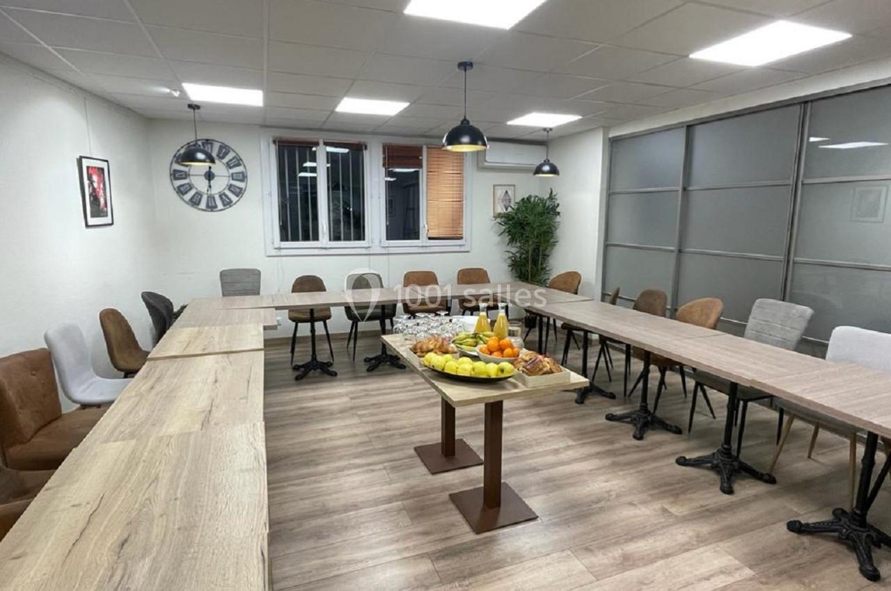 Salle de réunion lumineuse avec tables disposées en U, chaises variées et plateau de fruits au centre.