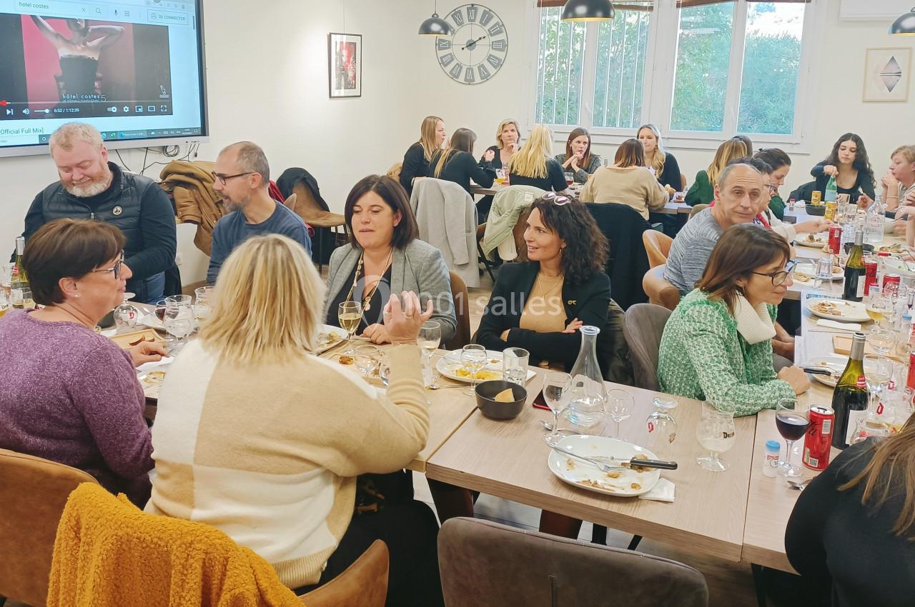 Des personnes partagent un repas convivial autour de grandes tables dans une salle lumineuse.