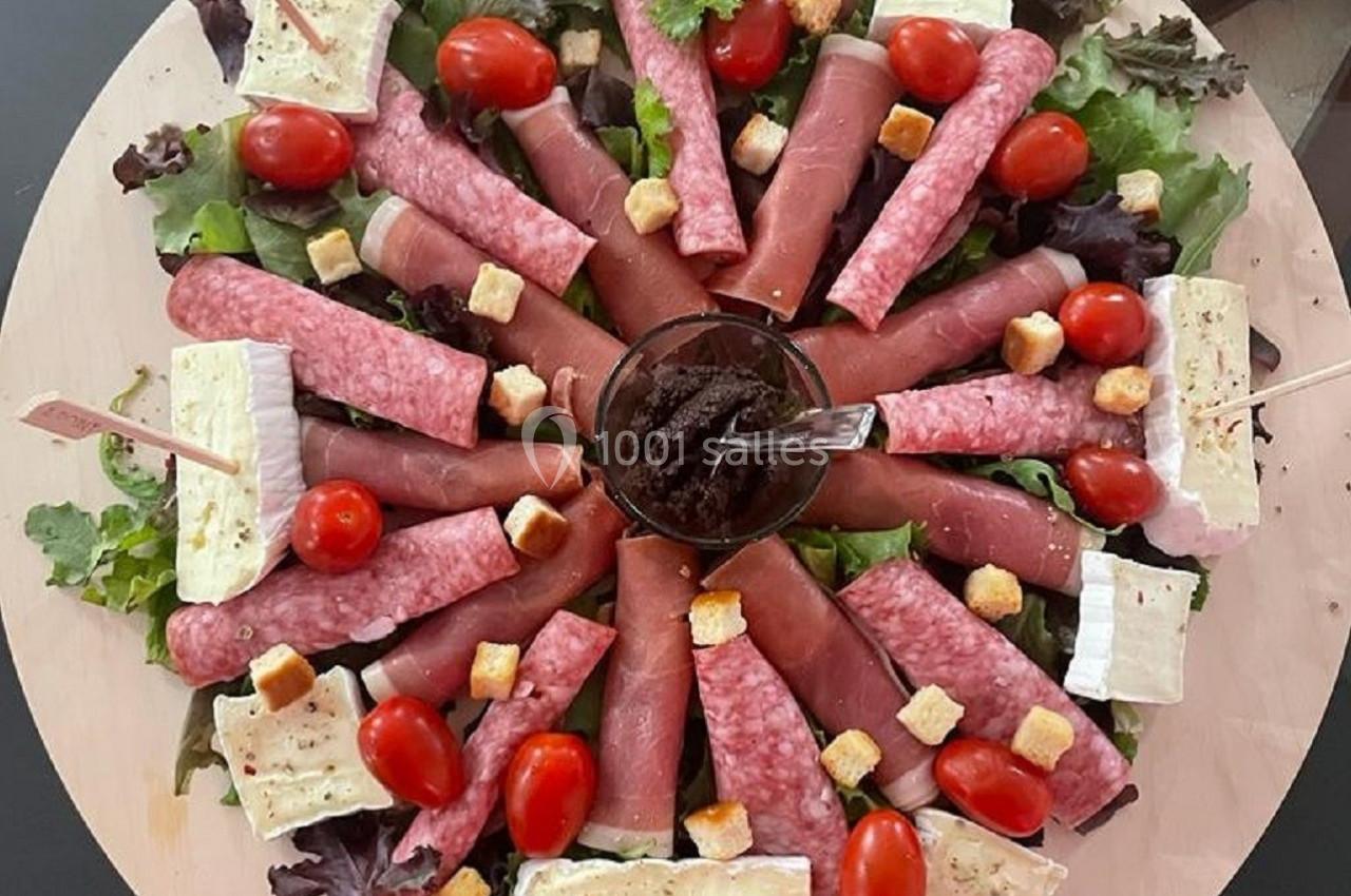 Plateau rond garni de charcuterie, fromage, tomates cerises, croûtons et salade, avec un bol de tapenade au centre.