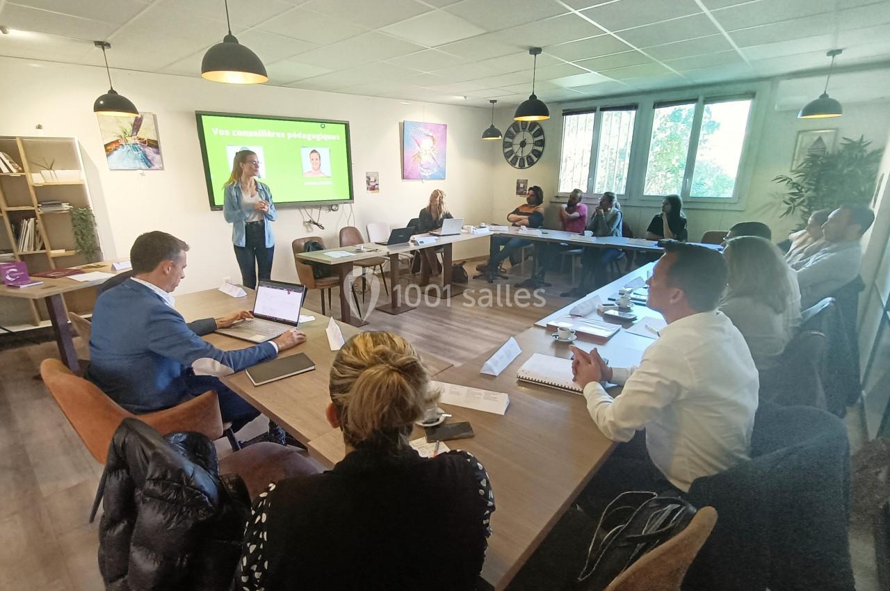 Une formatrice s'adresse à un groupe de personnes assises autour de tables dans une salle de réunion lumineuse.