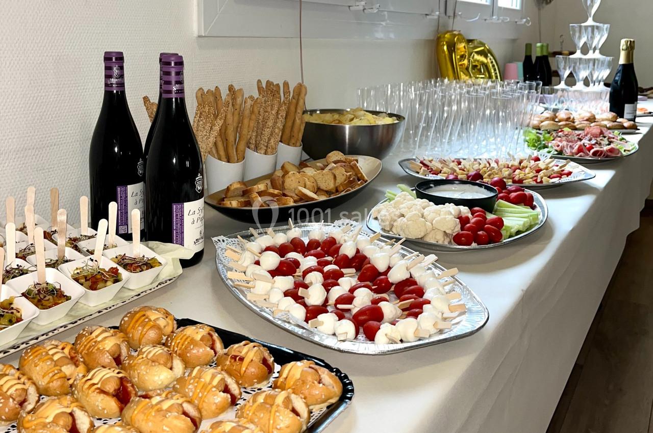 Buffet varié avec viennoiseries, brochettes, crudités, pains, bouteilles de vin et verres disposés sur une table blanche.