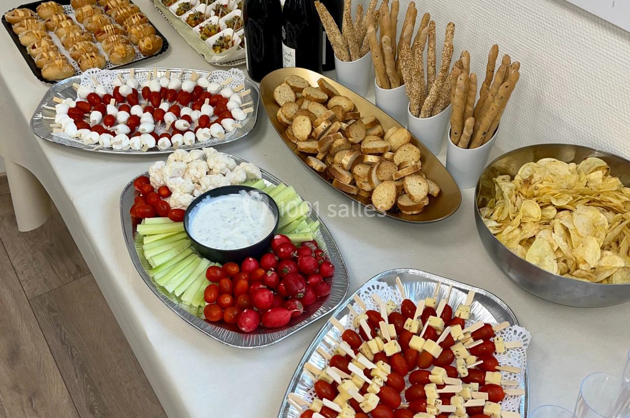 Buffet avec brochettes apéritives, légumes, chips, pains, verrines, et bouteilles de vin rouge sur une table.