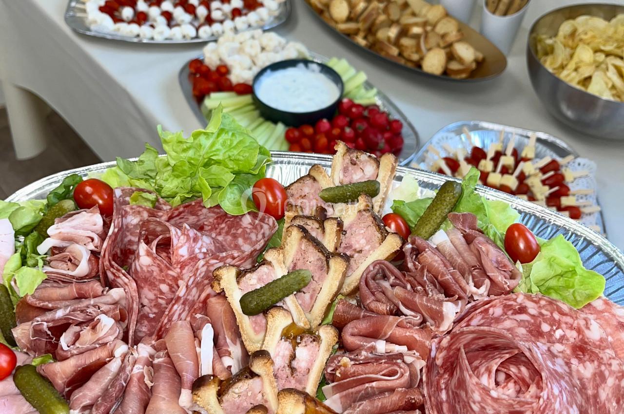 Plateau de charcuterie avec cornichons, tomates cerises et pâtés, entouré d'autres plats et bouteilles de vin en arrière…
