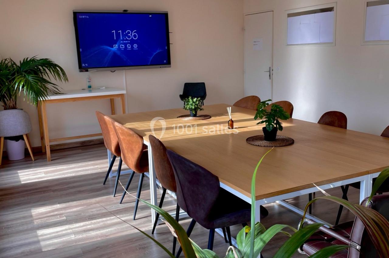 Salle de réunion lumineuse avec une grande table rectangulaire, des chaises, un écran mural et des plantes décoratives.