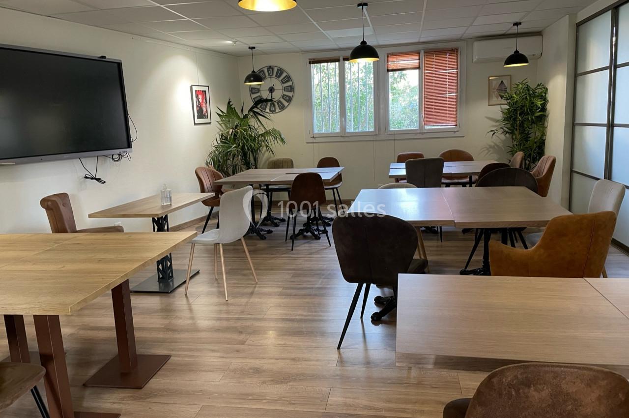 Salle lumineuse avec tables en bois, chaises variées, plantes vertes, tableau mural et grandes fenêtres.