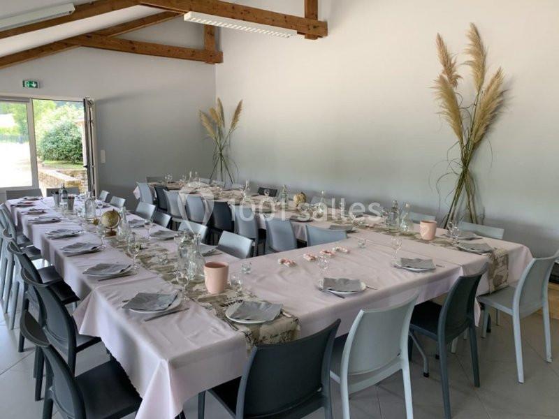 Salle décorée avec des tables alignées, nappes roses, chaises grises et ornements simples, prête pour un repas.