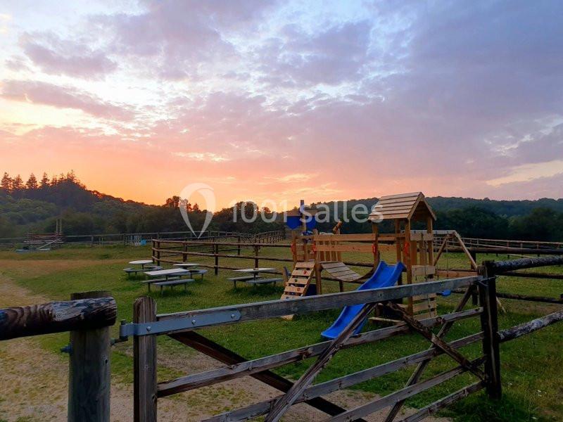 Aire de jeux en bois avec toboggan, entourée de clôtures, dans un paysage champêtre au coucher du soleil.