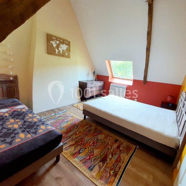 Chambre mansardée avec deux lits, tapis colorés, mur partiellement rouge et fenêtre de toit laissant entrer la lumière.