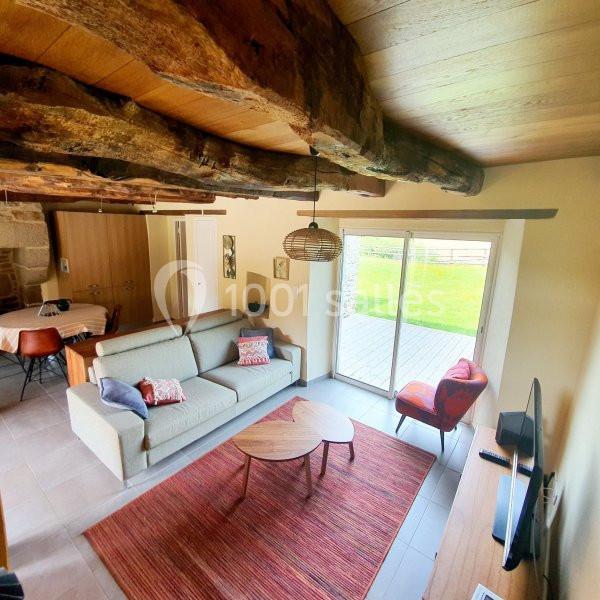 Salon lumineux avec poutres apparentes, canapé, table basse, tapis rouge, et baie vitrée donnant sur un jardin.