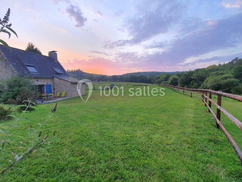 Maison en pierre avec toit en ardoise, entourée d'un grand champ verdoyant et d'une clôture en bois, au coucher du soleil.