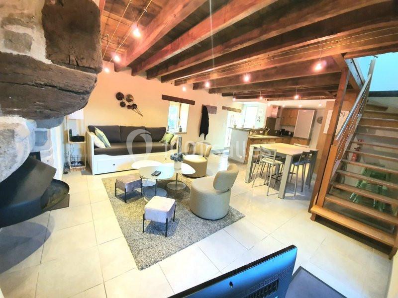 Salon lumineux avec poutres apparentes, canapé, fauteuils, table basse, cuisine ouverte et escalier en bois.