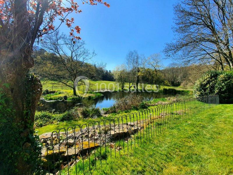 Paysage champêtre avec un petit étang entouré d'arbres, une pelouse et une clôture métallique au premier plan.