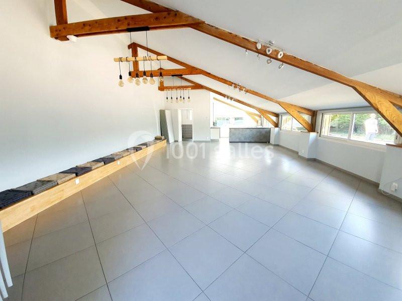 Salle lumineuse avec poutres apparentes, carrelage au sol, grandes fenêtres et éclairage suspendu moderne.