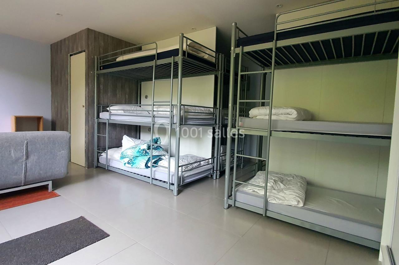 Dortoir avec lits superposés en métal, draps pliés, sol carrelé et un canapé gris dans un coin de la pièce.