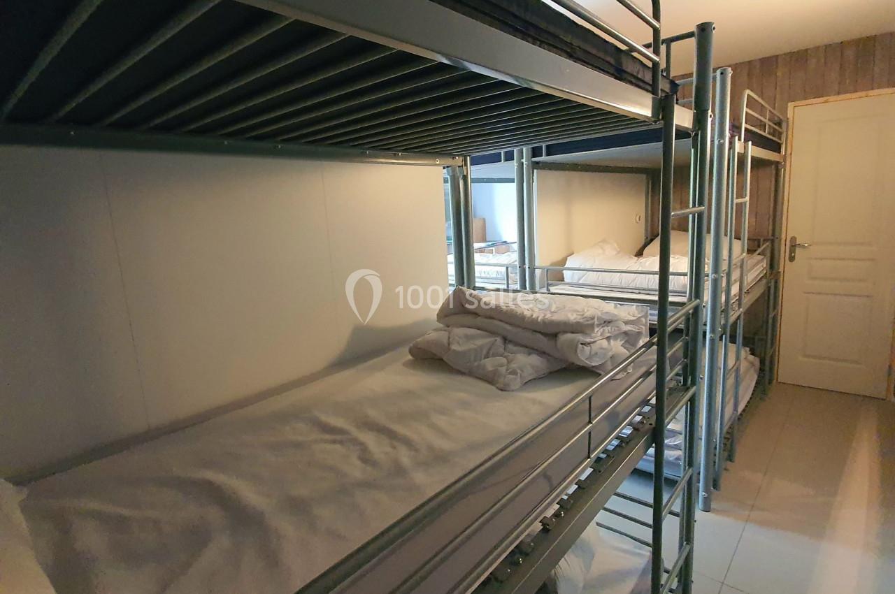 Dortoir avec lits superposés en métal, draps blancs pliés sur certains lits, sol carrelé et porte en arrière-plan.