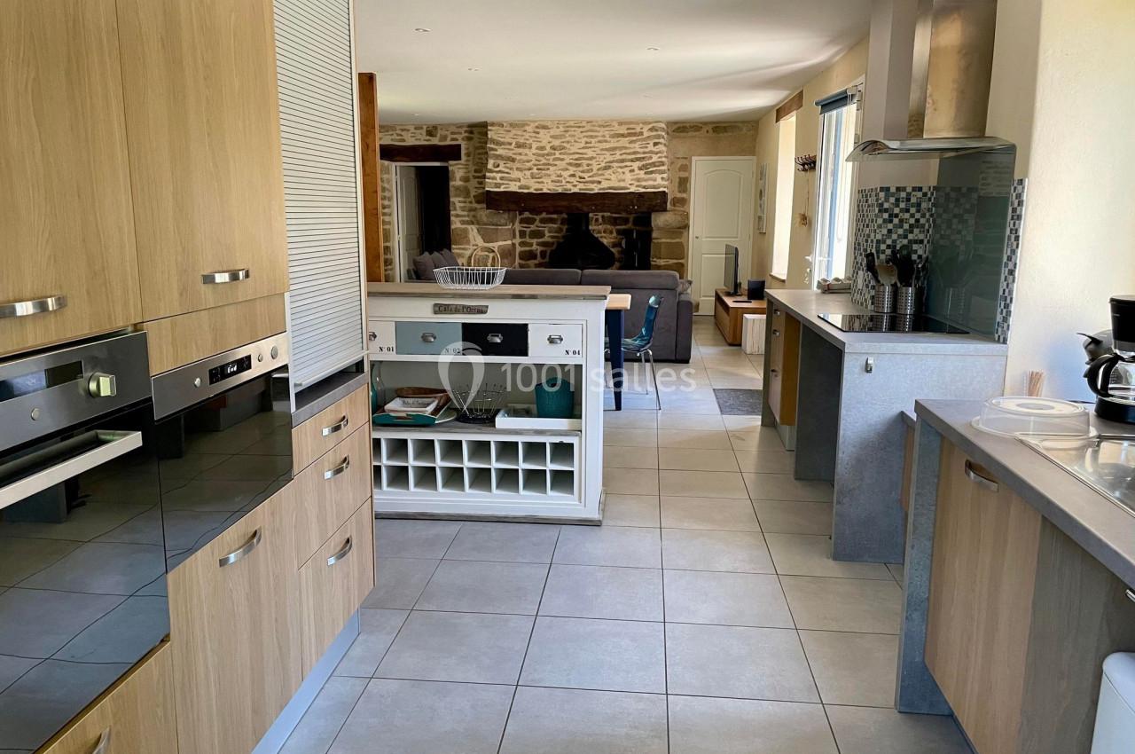 Cuisine moderne avec meubles en bois clair, plan de travail gris, électroménagers intégrés et vue sur un salon avec cheminée.