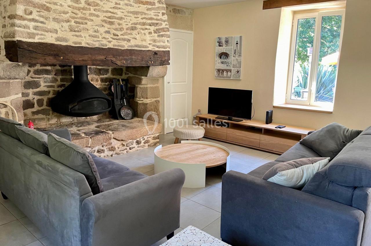 Salon avec cheminée en pierre, canapé gris, table basse ronde, télévision et fenêtre laissant entrer la lumière naturelle.