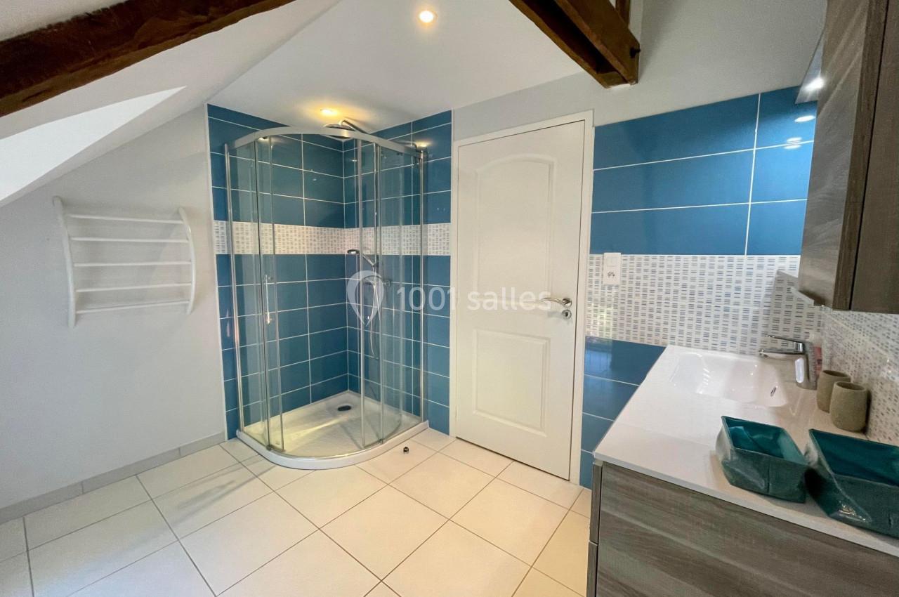 Salle de bain moderne avec douche à paroi vitrée, carrelage bleu, lavabo avec meuble et poutre apparente au plafond.