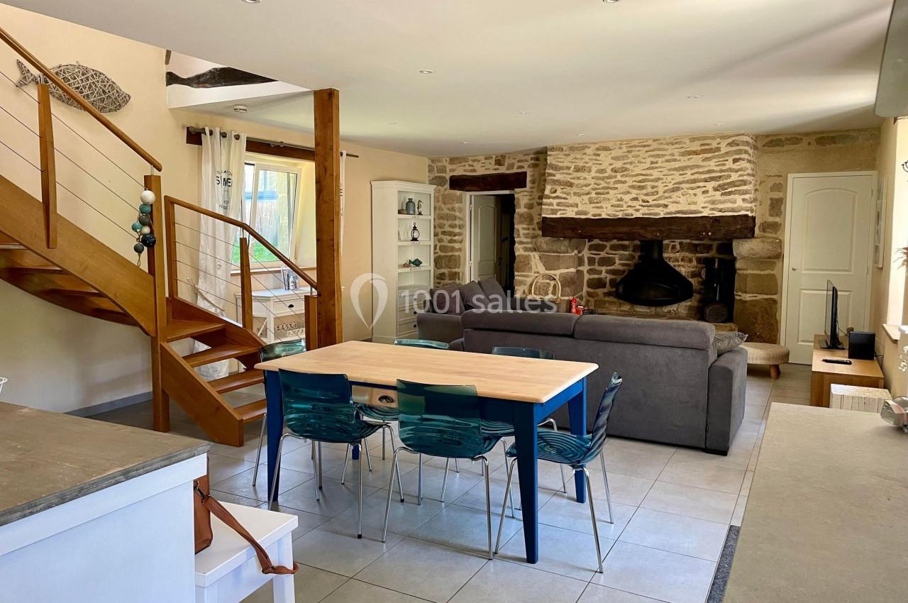 Salon lumineux avec murs en pierre, poêle à bois, escalier en bois, table à manger et canapé gris.