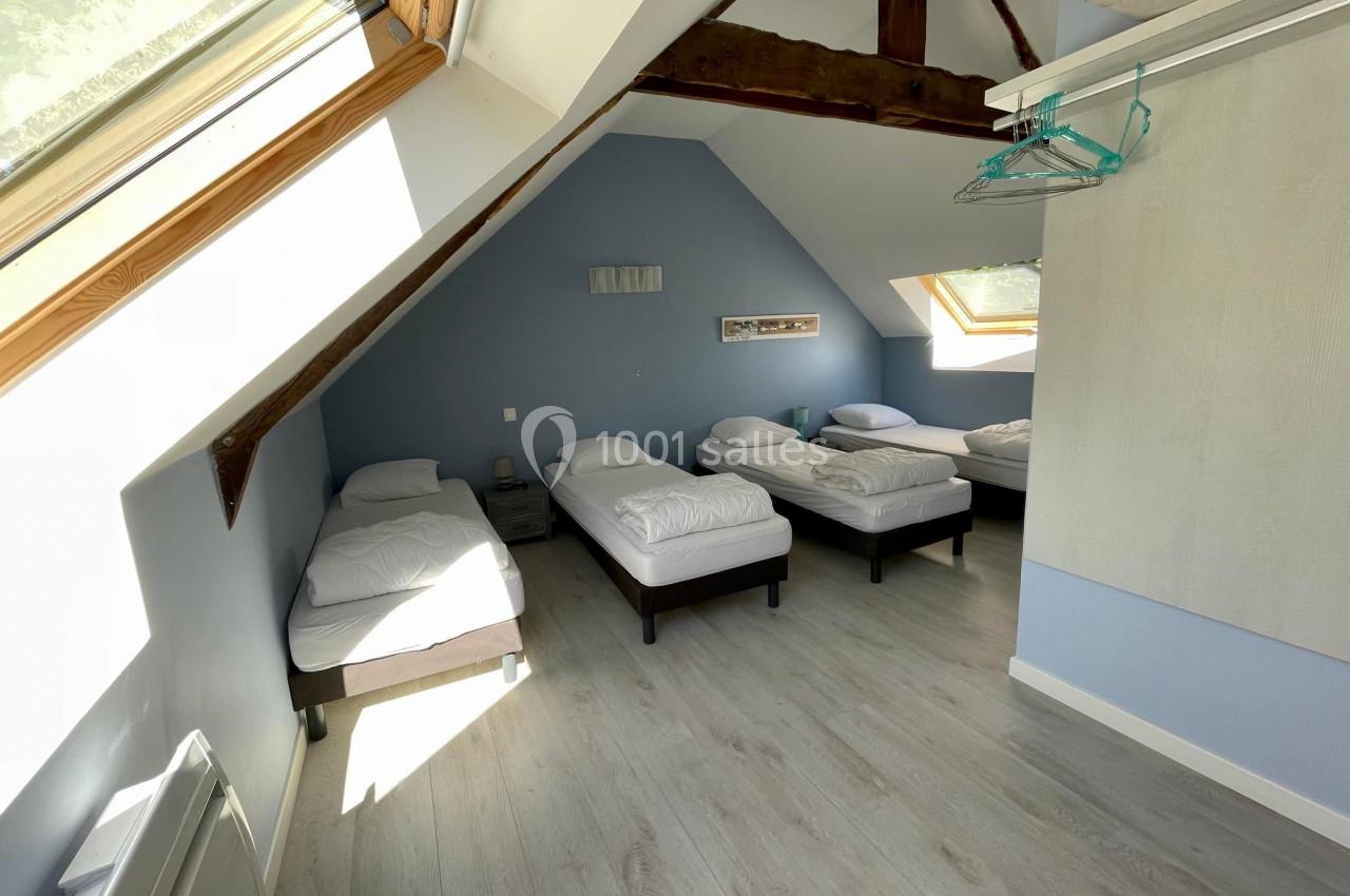 Chambre mansardée avec quatre lits simples, murs bleus, parquet clair et fenêtres de toit laissant entrer la lumière…