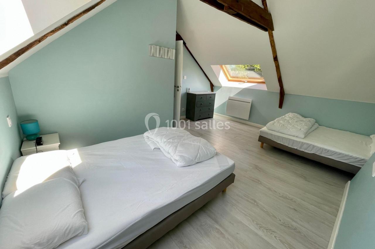 Chambre mansardée avec deux lits, parquet clair, murs bleus et poutres apparentes, éclairée par une fenêtre de toit.