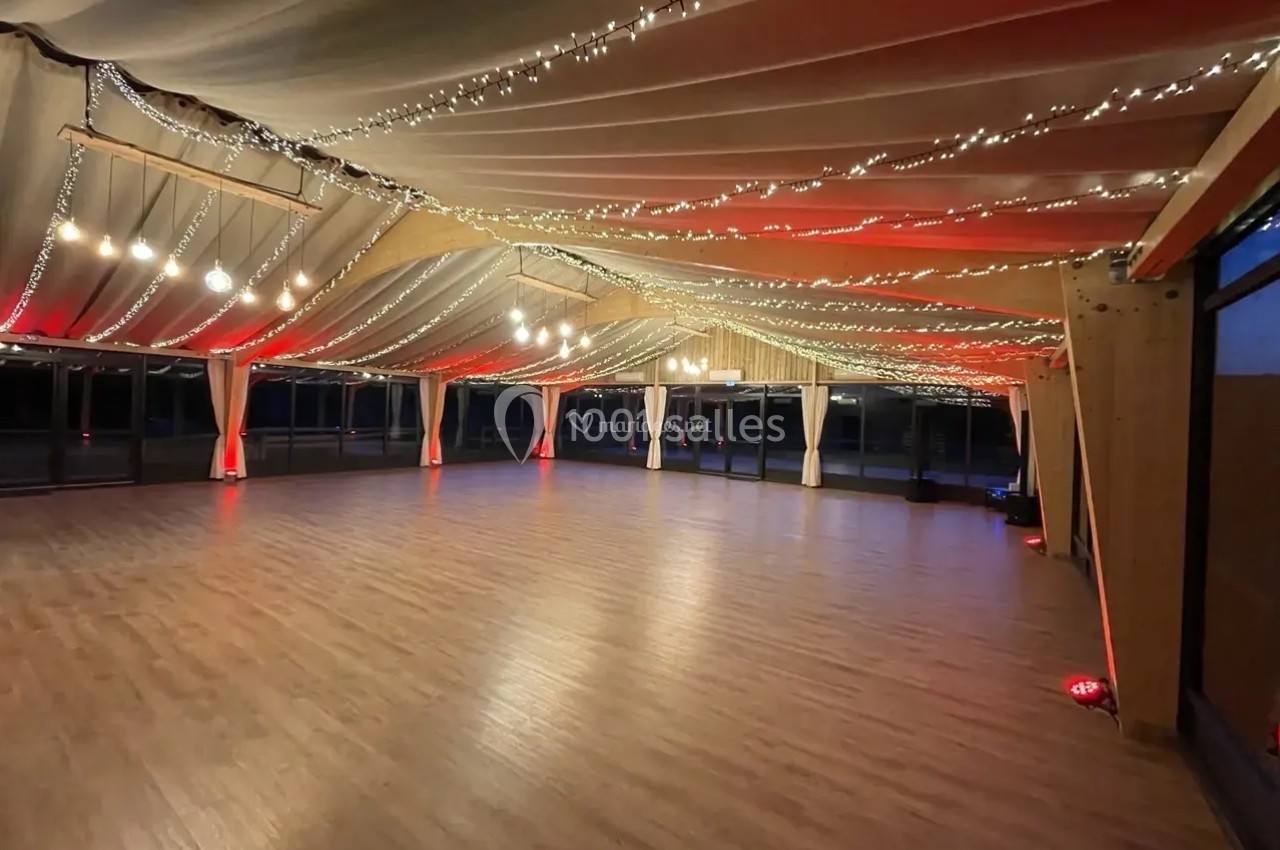Grande salle vide avec un parquet en bois, des tentures colorées au plafond et des guirlandes lumineuses suspendues.