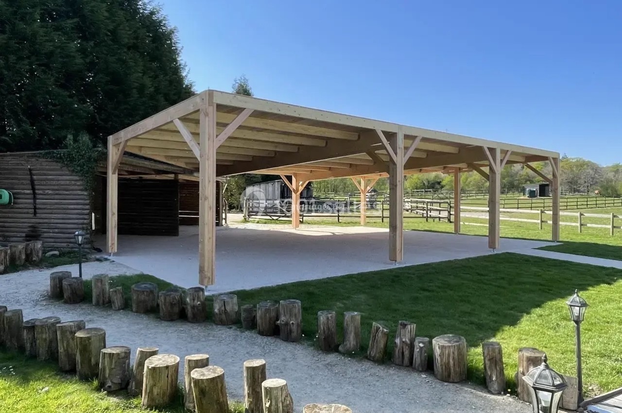 Structure en bois ouverte avec toit, située dans un espace extérieur verdoyant, entourée de rondins et de pelouse.