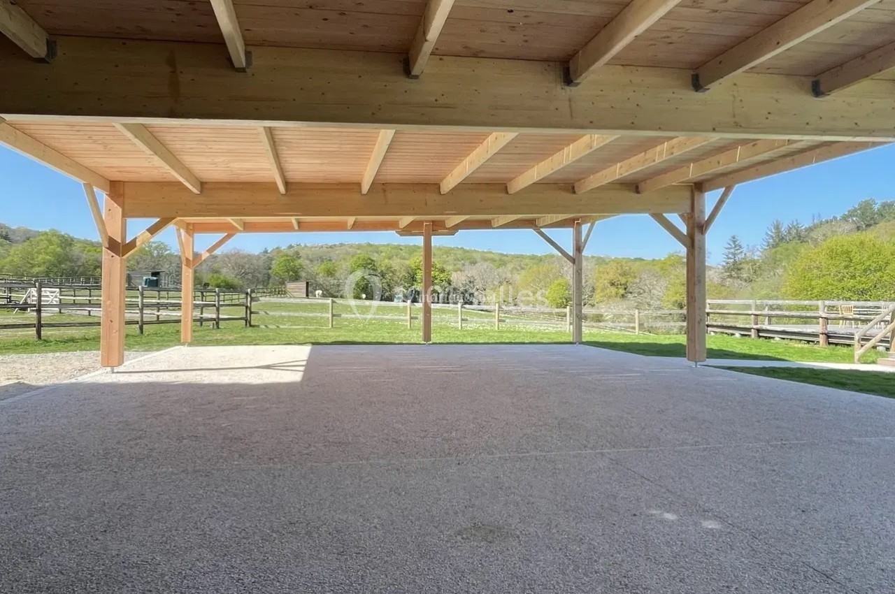 Structure en bois ouverte avec toit, offrant une vue sur un espace extérieur verdoyant et des clôtures en arrière-plan.