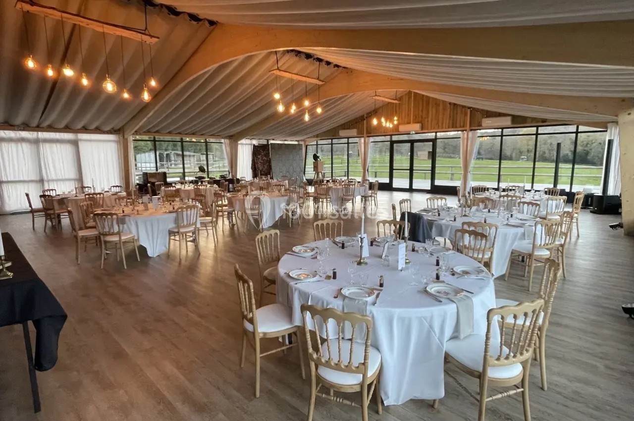 Salle de réception lumineuse avec tables rondes dressées, chaises en bois clair et grandes baies vitrées donnant sur l…