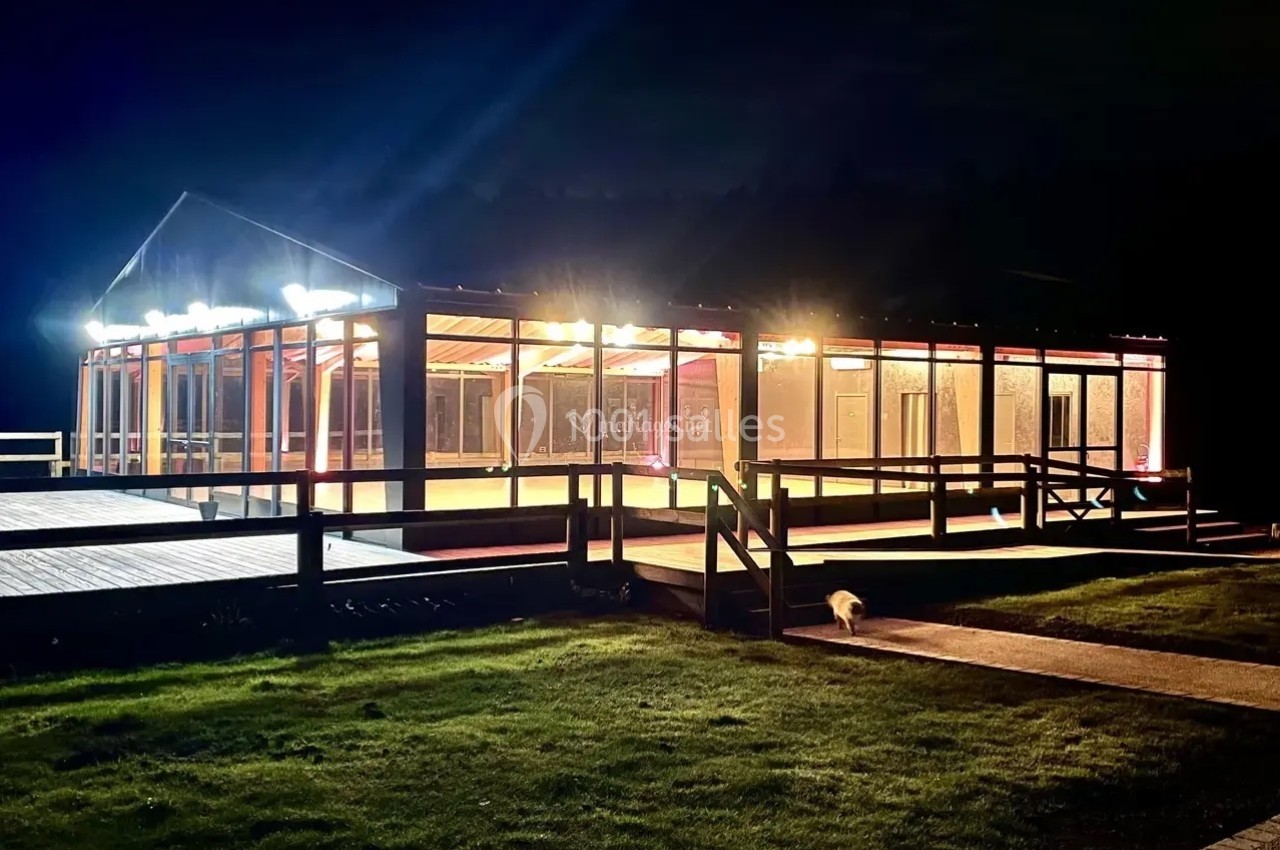 Bâtiment vitré éclairé de nuit, entouré d'une terrasse en bois et d'une pelouse.