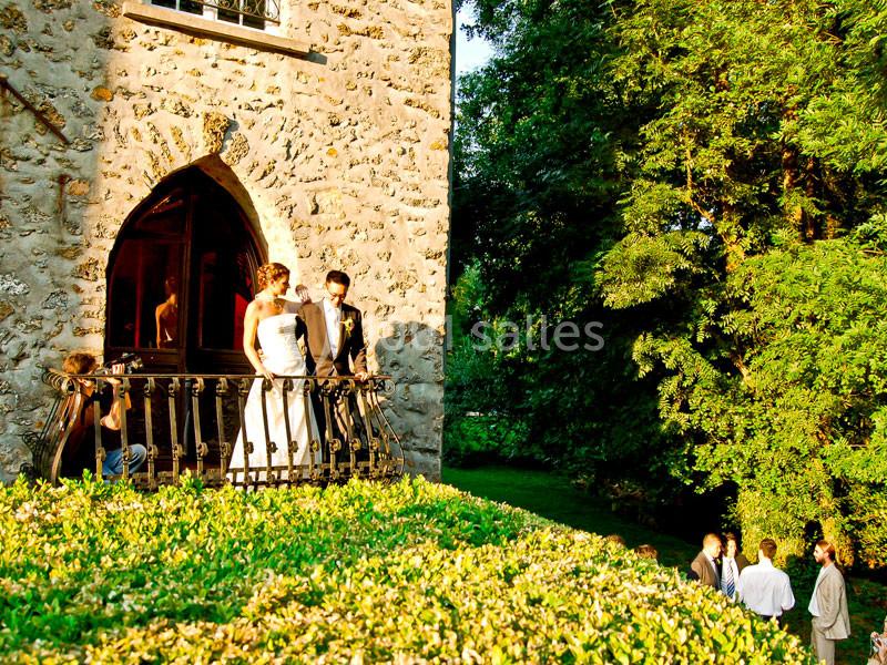 Un couple en tenue de mariage se tient sur un balcon en pierre, entouré d'invités et d'arbres verdoyants.