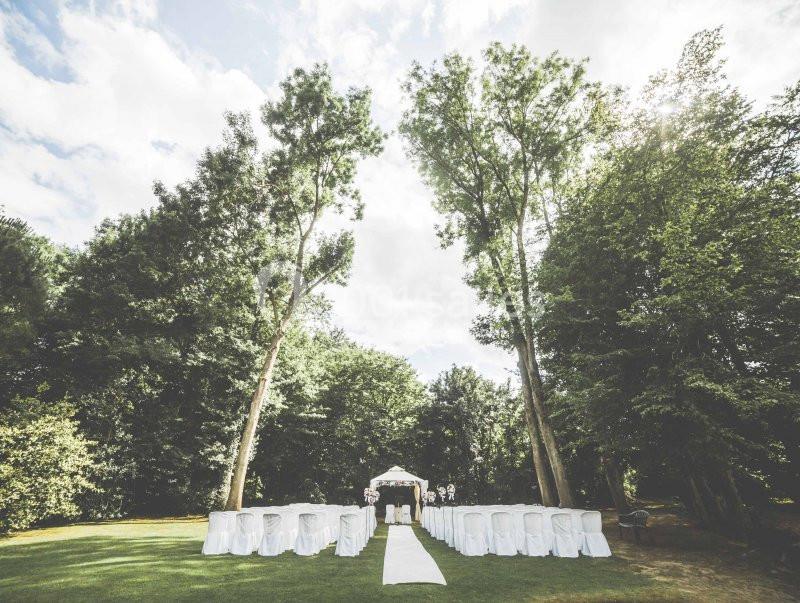 Chaises blanches disposées en rangées sur une pelouse, face à une arche décorée, entourées d'arbres sous un ciel…