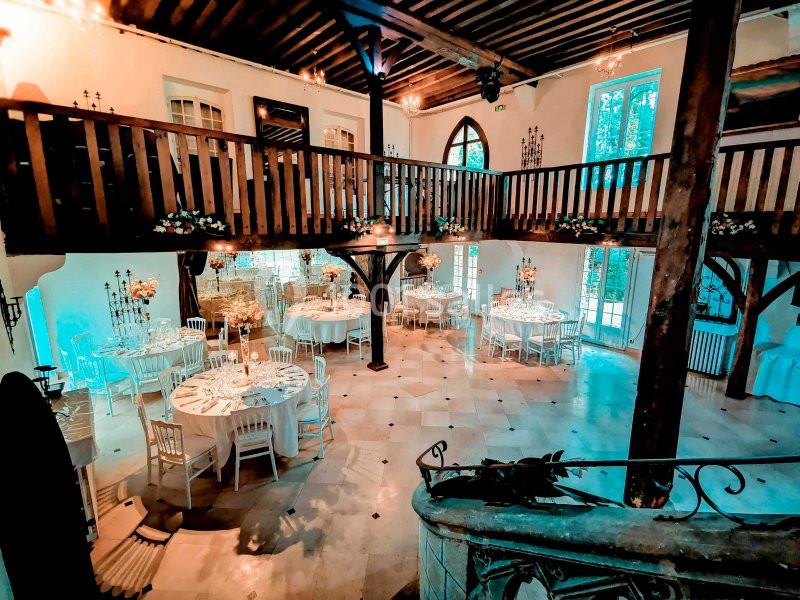 Salle de réception lumineuse avec mezzanine en bois, tables rondes dressées et décoration florale élégante.