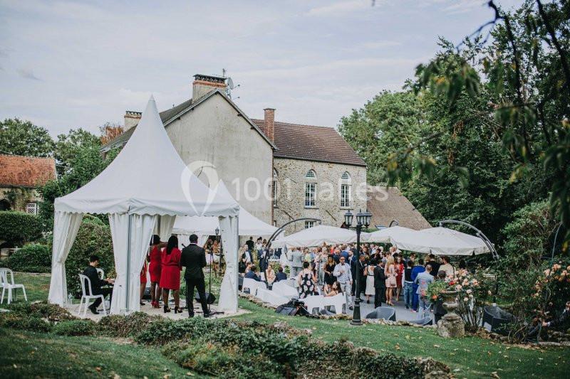 Des invités rassemblés sous des tentes blanches dans un jardin, près d'une maison ancienne, lors d'un événement en plein air.