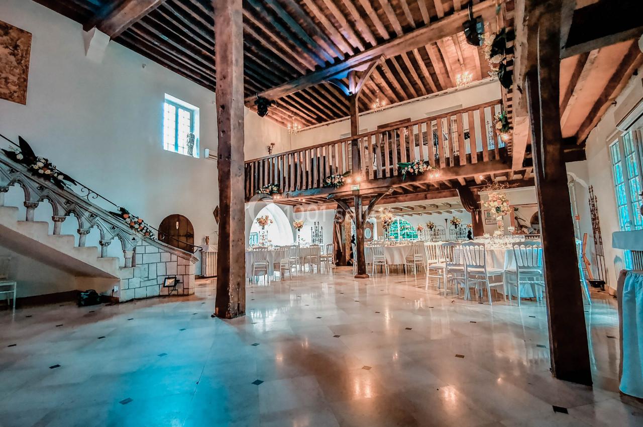 Salle de réception spacieuse avec poutres apparentes, mezzanine en bois, tables dressées et escalier décoré de fleurs.