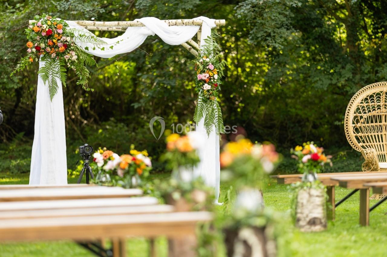 Arche de mariage décorée de fleurs et de tissu blanc dans un jardin, avec bancs en bois et chaise en osier.