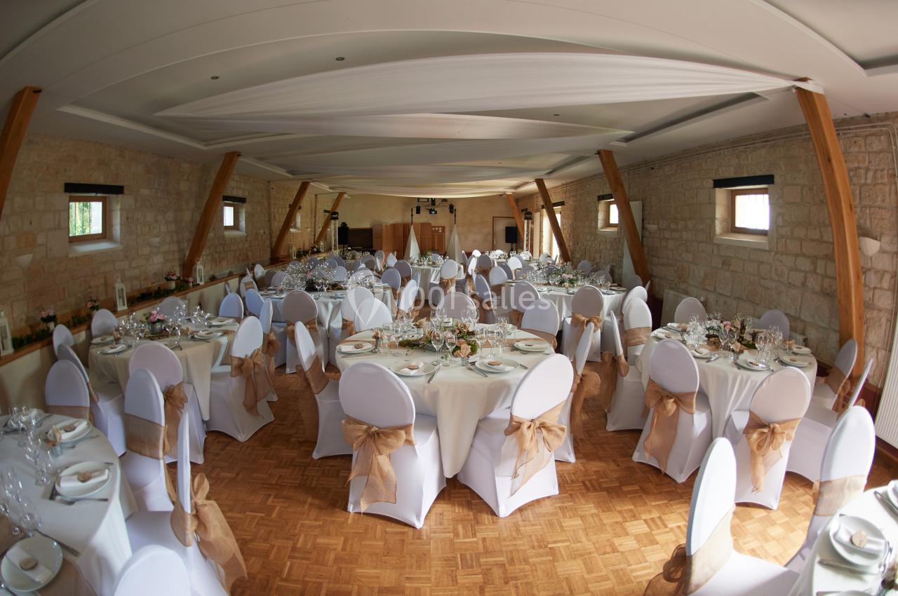Salle de réception décorée avec des tables rondes, nappes blanches et chaises ornées de nœuds en tissu beige.