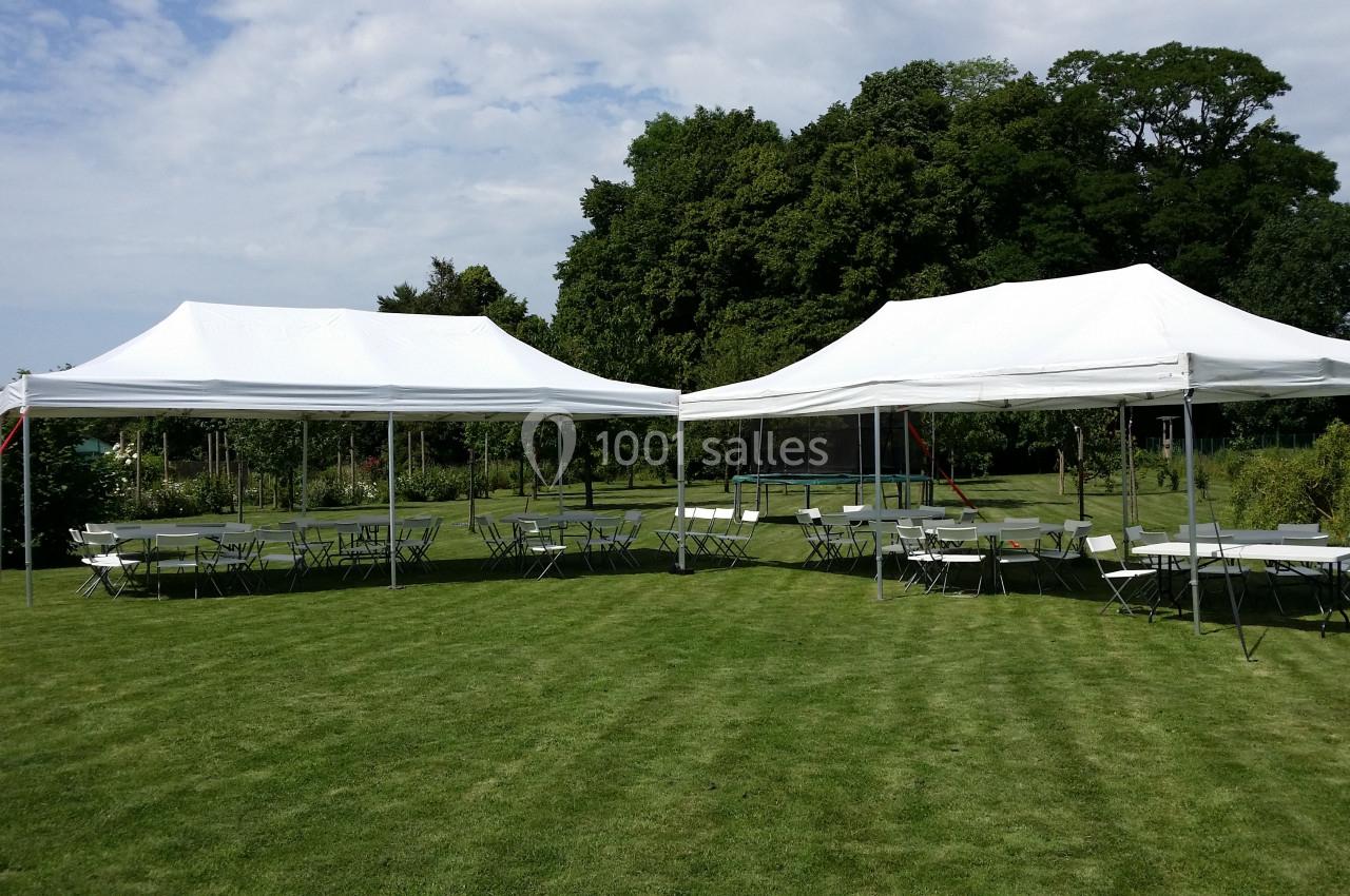 Deux tentes blanches montées sur une pelouse avec des tables et des chaises disposées autour, entourées de verdure.