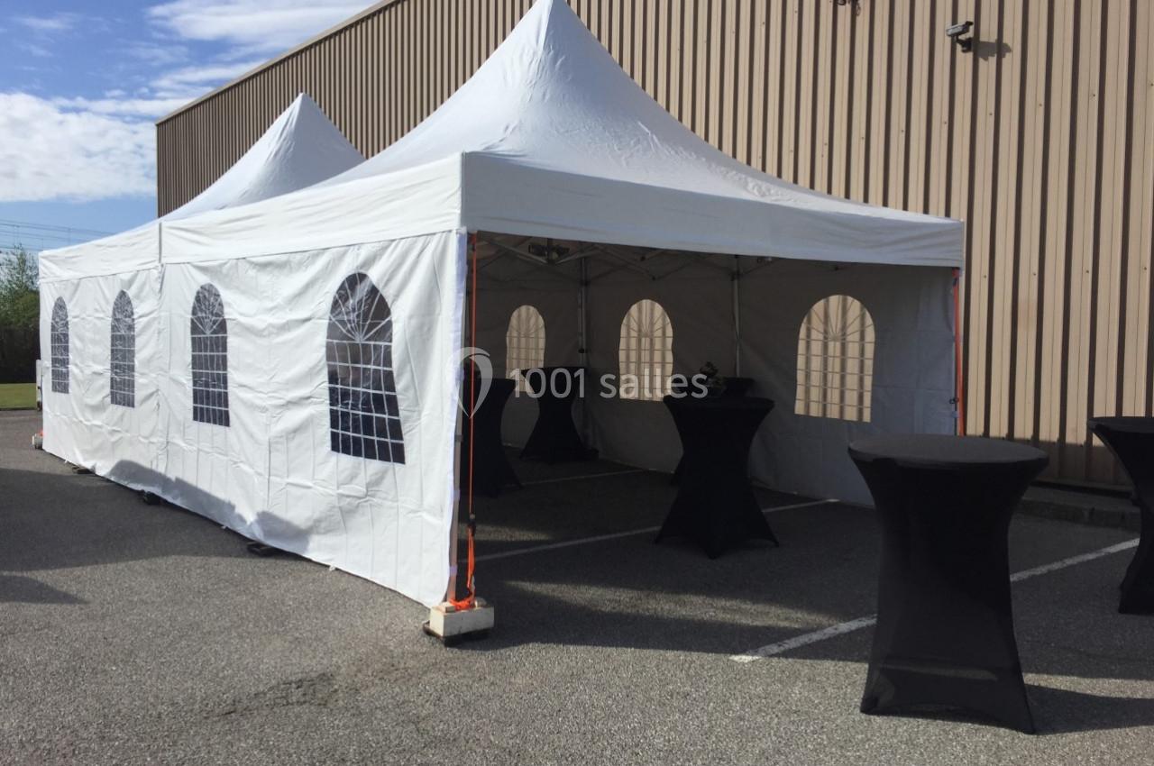 Tente blanche avec fenêtres transparentes installée sur un parking, entourée de tables hautes noires.