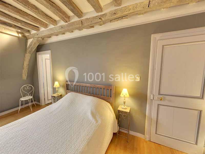 Chambre avec lit double, murs gris, poutres apparentes, deux lampes de chevet et une chaise en métal blanc.