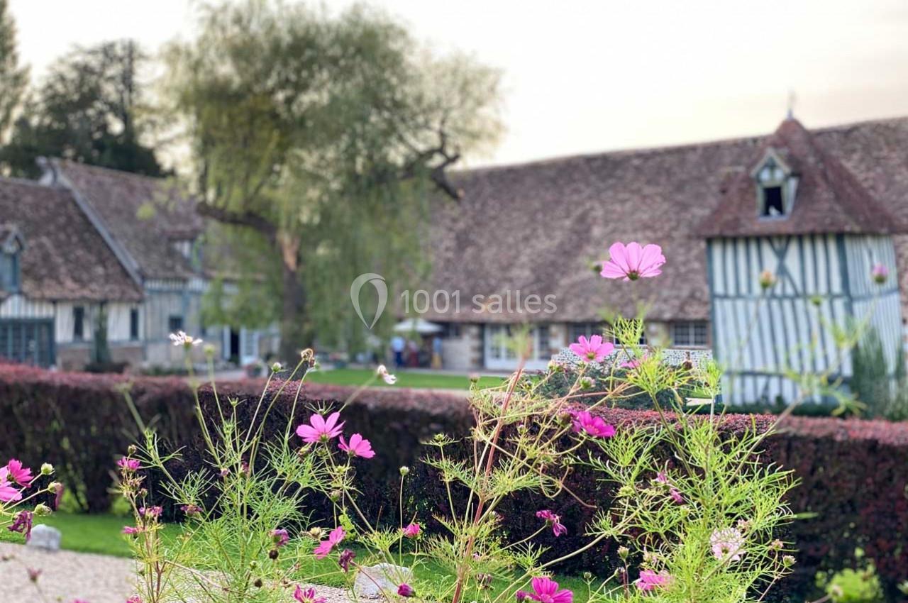 Jardin fleuri avec des cosmos roses en premier plan, devant des maisons à colombages et un arbre au fond.