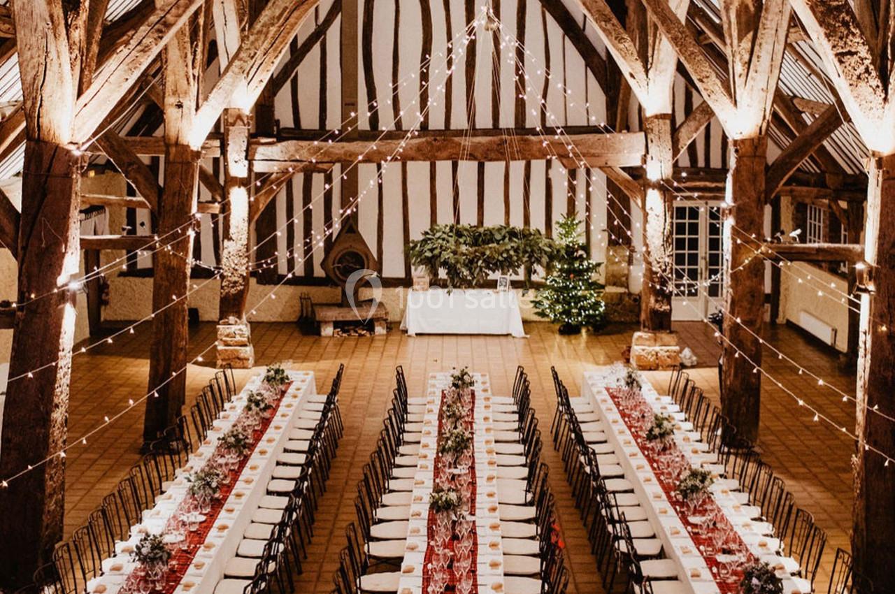 Grande salle de réception rustique avec poutres apparentes, tables longues décorées et guirlandes lumineuses suspendues.