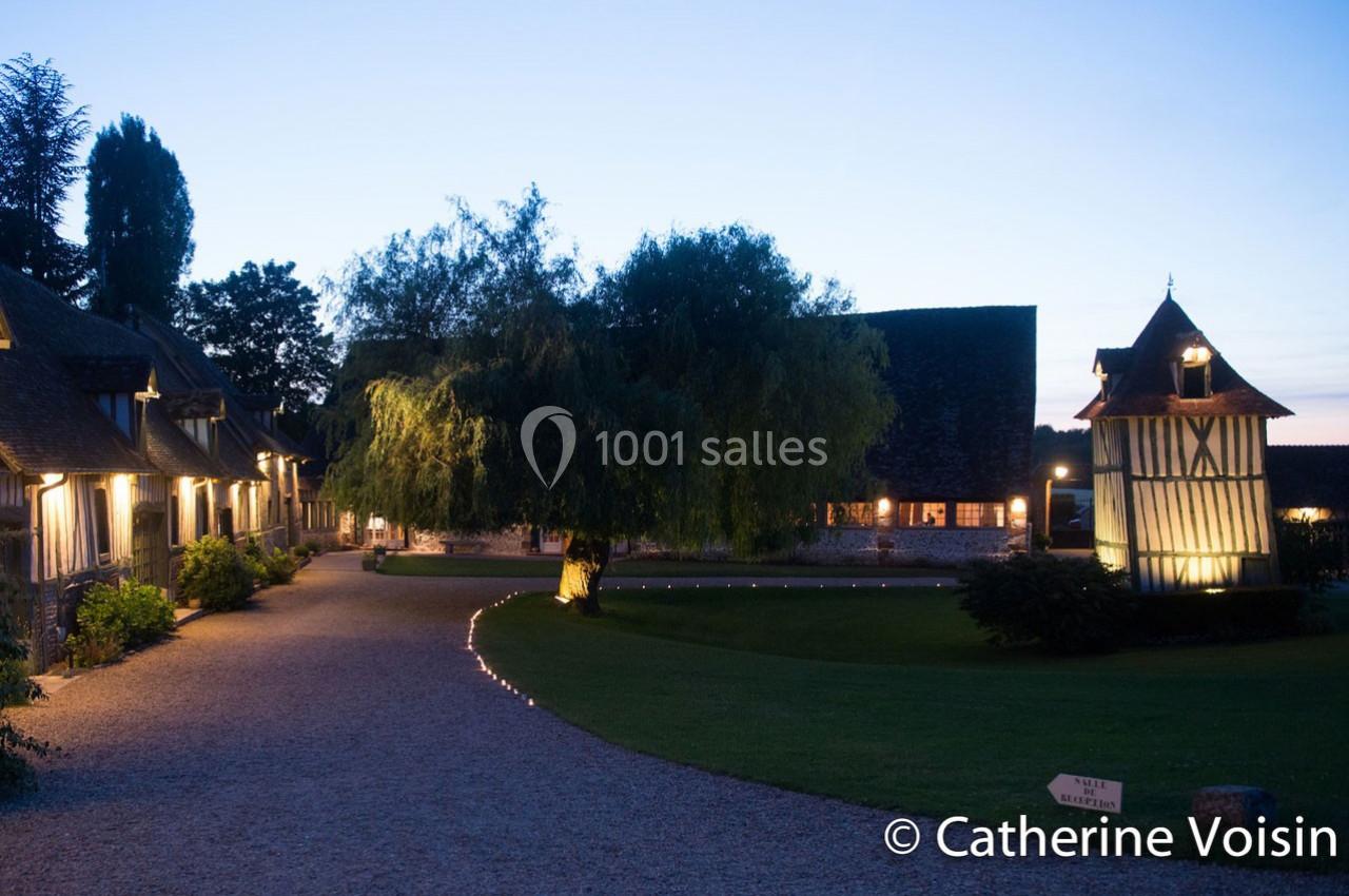 Chemin éclairé menant à des bâtiments traditionnels normands entourés de pelouses au crépuscule.