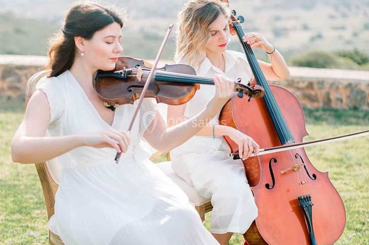 Deux musiciennes jouent du violon et du violoncelle en plein air, assises sur une pelouse sous un ciel dégagé.