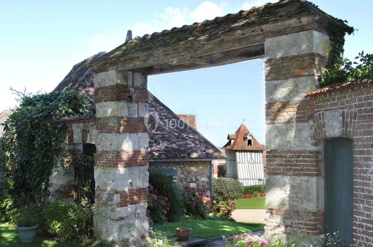 Porche en pierre et briques donnant sur une cour avec des bâtiments à colombages et un jardin fleuri.
