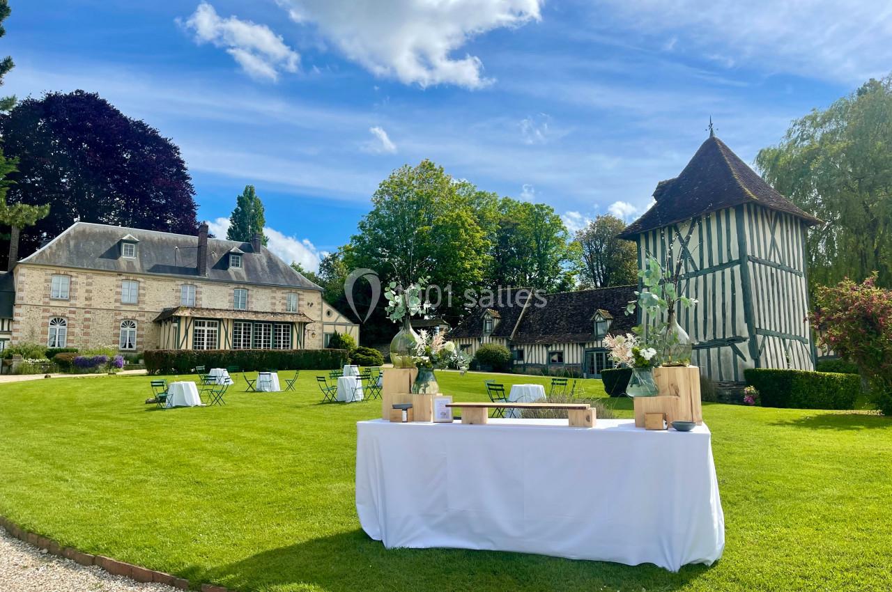 Jardin avec pelouse aménagée, tables et chaises, devant un manoir et une tour à colombages sous un ciel bleu.
