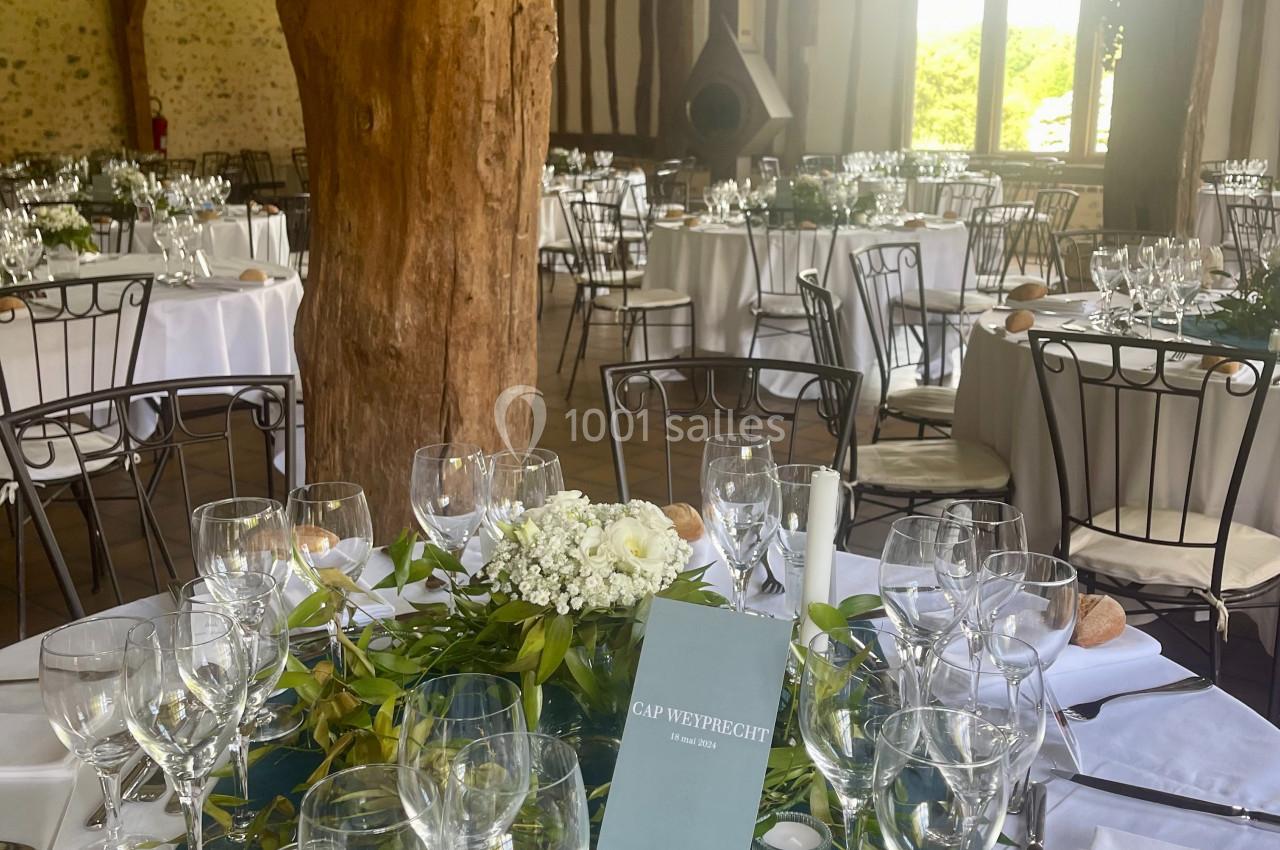 Salle de réception rustique avec tables dressées, nappes blanches, verres, couverts et décoration florale centrale.